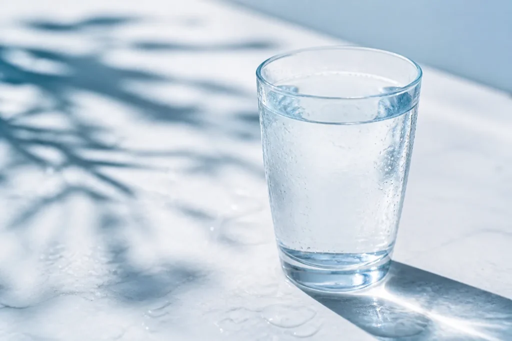 Create a clean, photorealistic editorial image featuring a single clear glass of water with fresh droplets on a bright, softly lit surface, subtle blu...
